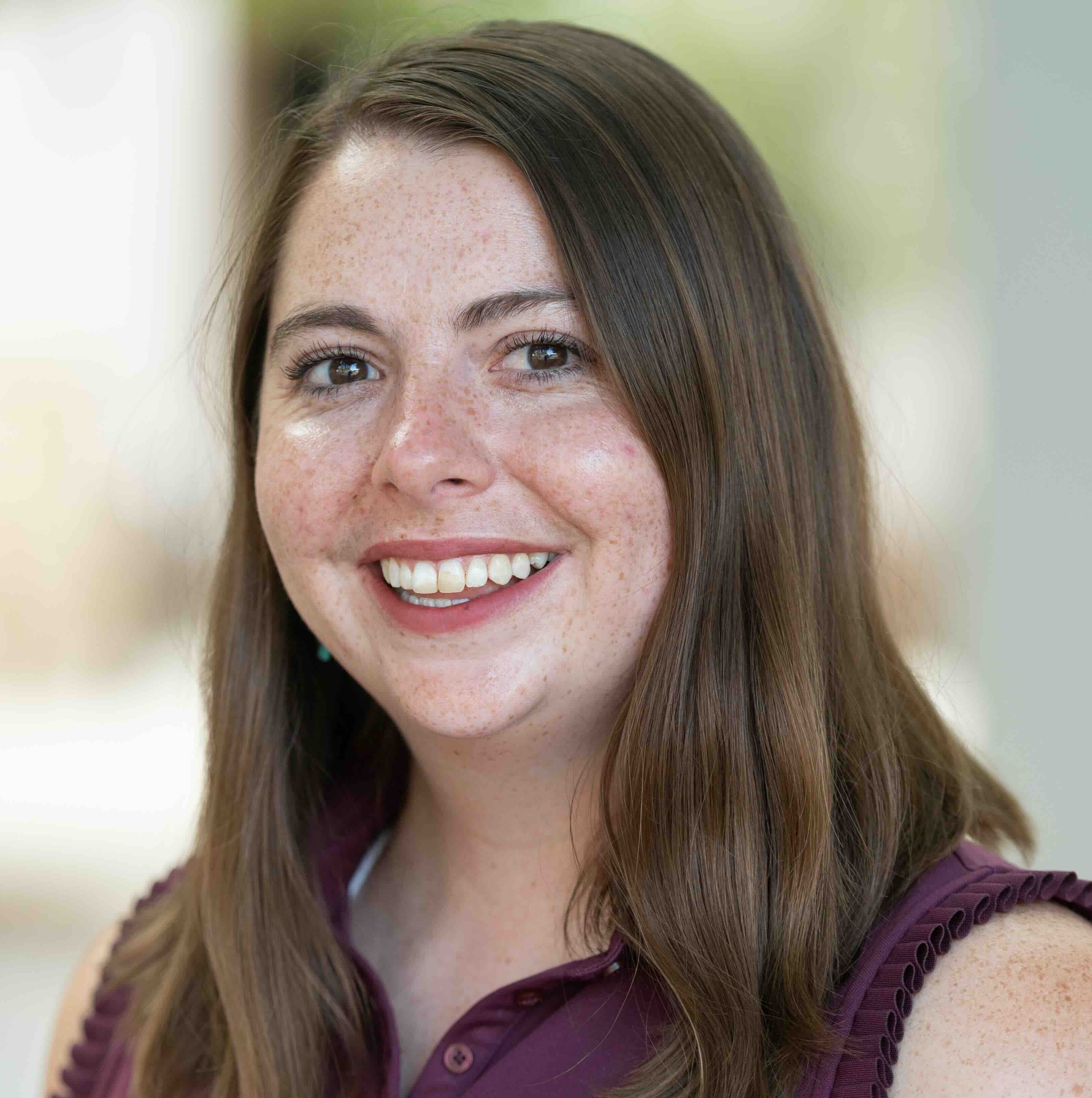 Smiling woman with long brown hair in a sleeveless purple top, photographed outdoors with a softly blurred background.